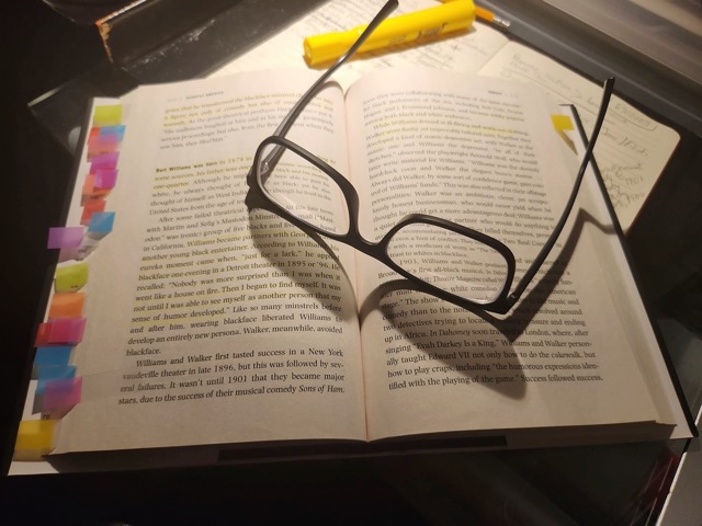 book open on a desk, littered with sticky notes, glasses hold the pages open